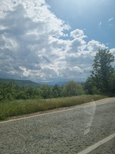 The image depicts a view from a roadside. The sky is largely filled with white, billowing clouds against a blue background, with bright sunlight in the upper left corner. A line of green trees and vegetation separates the sky from the gray asphalt road on the right side of the frame. The road surface appears worn. In the distance, a hint of mountains is visible, suggesting an elevated or rural location. The image captures a natural landscape along a roadway. The location is likely Хаджийско, Bulgaria.