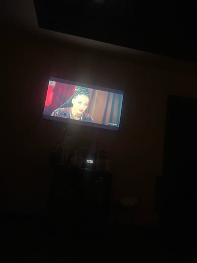 The image captures a dimly lit room where a television screen displays a woman with braided hair and makeup. She is wearing a patterned garment with red and blue accents. The backdrop on the screen appears to be a warm-toned, possibly wooden, setting. To the right of the woman on the screen, the time "22:50" is visible in white text. The television itself is mounted or placed on a surface, and some electronic equipment is visible below it in the dark. In the upper right quadrant of the image, a faint circular object, possibly a clock, is discernible against the dark wall. The overall scene suggests a private viewing of a show or movie in the evening. The context of "Приморский, Russia" provides a geographical location, but no specific environmental details like weather or outdoor cues are visible within the frame. The woman's gaze is directed away from the viewer, and her expression is somewhat intense, but emotions are difficult to ascertain definitively in this lighting. The text "LostFilm.TV" is also visible on the screen.