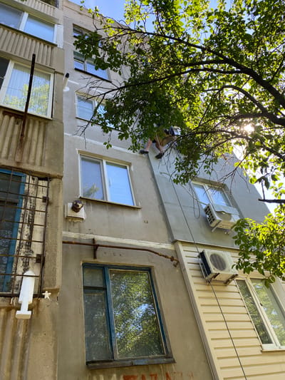 In the city of Zorynsk, Ukraine, an individual is seen rappelling down the exterior of a multi-story building, illuminated by bright sunlight filtering through lush green tree branches. The building's facade is a mixture of grey concrete and beige siding, with various windows, some featuring blue frames. Air conditioning units are mounted on the exterior walls. A metal grate is visible over one window on the left side of the building. The perspective is from below, looking upwards at the individual against the backdrop of the sky and foliage.
