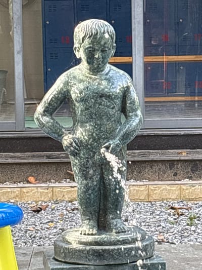 An outdoor forensic image depicts a stone statue of a nude young boy serving as a fountain, located in Ngũ Hành Sơn Ward, Vietnam. The statue, rendered in a speckled green and grey material, stands frontally with hands on hips, a stream of water emanating from its groin area and descending onto a circular, tiered stone base. In the immediate background, a structure with reflective glass panels is visible, reflecting multiple dark blue locker-like units. Some red numerical designations, such as '13', '16', '17', and '18', are discernible on or through these reflected units. Below this structure, a dark horizontal band rests above a light-colored, possibly tiled, wall. The ground around the statue consists of grey gravel with scattered dry brown leaves. A fragment of a bright blue and yellow plastic object is present in the lower left foreground. The scene is illuminated by natural light.