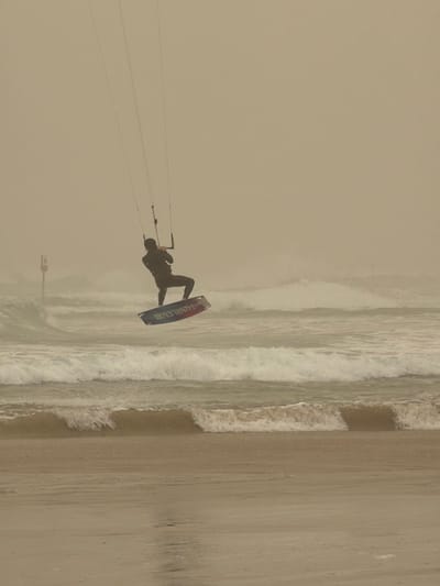 A kite surfer navigates choppy ocean waves along a sandy beach. The surfer, dressed in a dark wetsuit, is attached to a kite via lines and rides a blue and black board labeled "HARLEVE". The scene is overcast, creating a monochromatic, muted tone. Waves break close to the shoreline, marked by a distinct line where the wet sand meets the dry. In the background, a small, partially obscured structure stands on the beach.