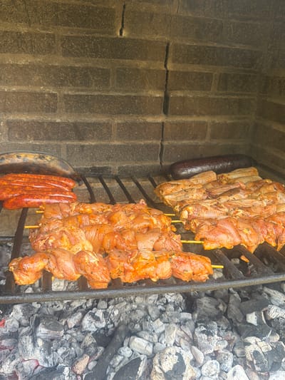 The image shows food grilling on a barbecue. In the foreground, skewers of marinated chicken are arranged on a black metal grill. Behind the chicken, several lengths of red sausages are laid out. Further back, a dark, elongated sausage or blood sausage is visible, along with what appears to be a grilled plantain. The barbecue is built with rough, reddish-brown bricks, and the grill is positioned over a bed of glowing charcoal. The scene suggests an outdoor cooking event.
