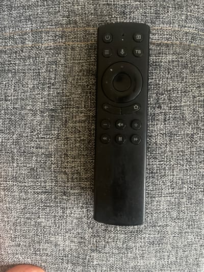 This media file features a black remote control lying on a textured gray fabric surface. The remote has several buttons, including a power button (represented by a circle with a vertical line), a home button (represented by a house icon), a back button (represented by an arrow pointing left), and navigation buttons arranged in a circular pattern. There are also buttons for volume control (plus and minus symbols) and playback control (play/pause and rewind/fast forward symbols). The fabric surface appears to be upholstery, possibly from a couch or chair, with a woven pattern of black, white, and gray threads. No people or any other objects are visible in the frame. The lighting suggests an indoor setting, with no indication of the time of day or weather conditions. There is no visible text on the remote control, other than the icons representing button functions. The scene is static, with no activity taking place. The location context provided is Berd, Armenia, but there are no visual cues within the image to confirm this specific location.