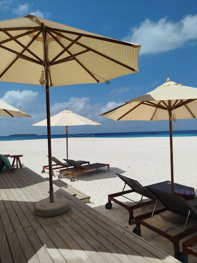 The image depicts a serene beach scene in Kendhoo, Maldives, under a bright blue sky with scattered white clouds. The foreground is dominated by a wooden deck, with a large, striped umbrella providing shade. Several lounge chairs, some with dark fabric and wheeled frames, are placed on the white sand. In the midground, two more umbrellas are visible, along with a volleyball net stretched across the beach. The pristine white sand slopes gently towards the turquoise ocean, which meets the horizon with a faint line of land in the distance. The water is calm and reflects the blue of the sky. The overall atmosphere is one of tranquility and relaxation, typical of a tropical island resort.