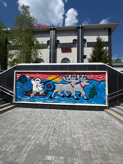 The image depicts an outdoor scene in the city of Neryungri, Russia. In the foreground, a paved walkway made of grey interlocking pavers leads up to a colorful bas-relief mural. The mural is set within a black frame and features a stylized depiction of a person in a white hooded suit on a sled, being pulled by two reindeer. The background of the mural shows a vibrant landscape with a setting sun, red waves, green trees, and blue water. Above the mural, the facade of a building is visible. The building has a modern design with horizontal bands of light and dark grey material, arched windows, and a prominent digital clock displaying "16:06". Red lettering, partially obscured by a tree, is also visible on the upper part of the building. The sky is blue with scattered white clouds. The scene is illuminated by daylight, suggesting it is daytime. The overall impression is of a public space adorned with art, possibly a transportation hub or a public building in Neryungri.