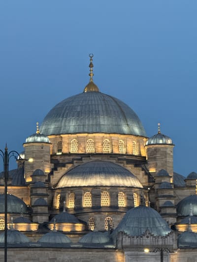 The image shows a close-up view of the Yeni Cami (New Mosque) in Fatih, Istanbul, Turkey, at dusk. The mosque's grand central dome and several smaller domes are visible, illuminated from within, casting a warm glow. The architectural details of the mosque, including its stone facade and arched windows, are prominent. The sky is a deep, clear blue, typical of twilight. A street lamp with a decorative wrought iron pole is partially visible in the foreground on the left. No people are present in the image. The focus is on the impressive structure of the mosque against the darkening sky.