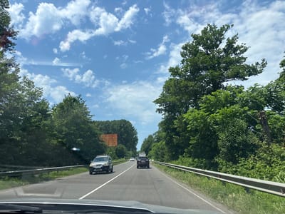 The image captures a view from inside a vehicle traveling on a two-lane road surrounded by lush green trees. The sky above is a bright blue, dotted with fluffy white clouds. Several vehicles are visible on the road, including a silver SUV in the foreground on the left, a dark red SUV further ahead, and a white van in the distance. On the left side of the road, a metallic guardrail runs along the edge, bordered by grass and dense foliage. On the right, a similar guardrail is present, also with abundant greenery. In the distance, a large, empty orange billboard is visible on the left side of the road, partially obscured by trees. The overall scene depicts a scenic drive through a verdant area.