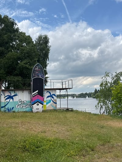 The image depicts a waterfront scene in Chiveryovo, Russia, under a partly cloudy sky. In the foreground, a grassy embankment slopes down towards a body of water. A stand-up paddleboard, decorated with abstract colorful designs and the "IBOARD" logo, is propped upright against a white, graffiti-adorned wall. The graffiti features stylized palm trees and the text "Can бордис". Behind the paddleboard and the wall, a metal platform structure with railings extends outwards.

The body of water, likely a lake or river, stretches across the mid-ground, reflecting the sky. Several small sailboats are visible on the water in the distance, along with a treeline and buildings on the opposite shore. Lush green trees frame the scene, with one prominent birch tree with white bark on the right side of the image. The overall atmosphere is that of a recreational area by the water.