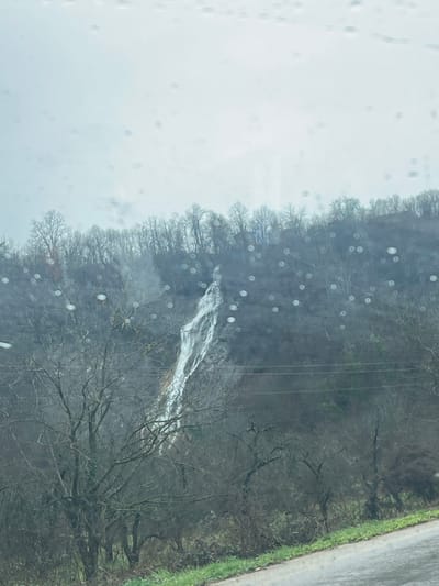 The image depicts a wooded hillside with a waterfall or rapid visible through bare trees. The foreground of the image is blurred with water droplets on a window, suggesting the photo was taken from inside a vehicle. The sky appears overcast and gray. A road borders the woods on the right side of the image. No people, subjects or actions, or interactions are visible. The location in the city of Касапите, Bulgaria is unknown.