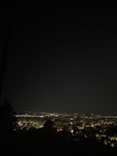An aerial night view of the city of Xanthi, Greece, is captured from a hillside overlooking the urban landscape. The city lights create a warm, diffused glow against the dark sky, illuminating the dense network of buildings. The foreground is dominated by the dark silhouettes of trees and foliage, framing the view of the city below. The perspective suggests the photograph was taken from a vantage point at a significant elevation. The lights are a mix of yellow and white, indicating streetlights and illuminated windows. The overall impression is a serene and sprawling urban scene at night.