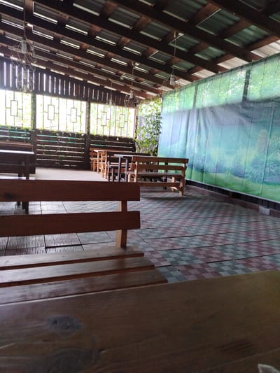 An outdoor seating area under a wooden roof is depicted. The area is furnished with several wooden benches and tables. The floor is covered with square tiles arranged in a checkered pattern of red and grey. In the background, a large green tarp with a printed landscape scene hangs on the right, and windows with wooden shutters are visible on the left, with greenery growing around them. Several decorative light fixtures hang from the ceiling. The scene suggests a public or semi-public space, possibly a cafe or restaurant patio, located in the settlement of Zyryanskoye, Russia.