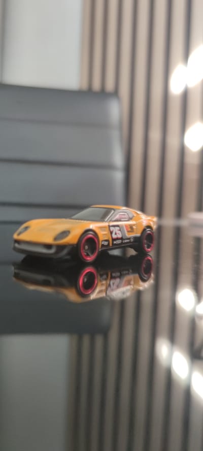 A yellow toy car with red rims is positioned on a reflective black surface. The car features the number 25 and other racing decals. The background consists of vertical blinds or curtains in muted tones with bright, out-of-focus lights. The scene appears to be indoors, possibly in a living room or office setting, and is not located in Liebenburg, Germany.