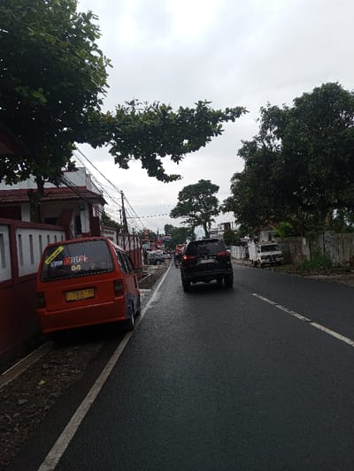 The image depicts a street scene under an overcast sky in Kamji Na-Yekha, Bhutan, with a wet asphalt road surface. In the foreground on the left, an orange-red minivan, identified by its yellow license plate "F 1916 TU" and "04" sticker, is parked on an unpaved shoulder next to a red-trimmed concrete wall belonging to a building with a reddish roof. Overhead, dense green foliage from a large tree and multiple electrical wires are visible. On the wet two-lane road, a dark-colored SUV is observed driving away from the viewer in the right lane. Further down the street, additional vehicle traffic is present. On the right side of the road, a large tree with dark green foliage stands prominently, followed by a light-colored utility truck parked on the shoulder next to a worn concrete wall. No individuals are discernible in the scene.