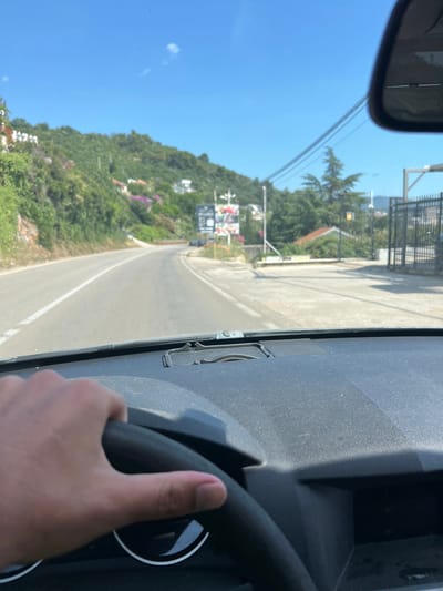 The image is taken from the interior of a vehicle, showing a hand gripping the steering wheel. The view is through the windshield, looking forward onto a paved road that curves to the left. The road is bordered by lush green vegetation on the left, with some white buildings visible on the hillside. On the right side of the road, there are trees, a dark metal fence, and some structures that appear to be commercial. In the distance, there are more hills and buildings. The sky is clear and blue with a few small white clouds. The scene appears to be a sunny day in a coastal or hilly region, likely Dobra Voda, Montenegro, given the presence of Mediterranean vegetation and architecture.