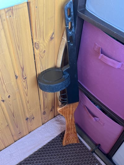 The image shows a close-up of a wooden-paneled wall and a dark-colored object with a drum magazine, resembling a firearm accessory, leaning against it. To the right of the object, part of a shelving unit with purple fabric storage bins is visible. A light-colored, textured mat and a piece of pink fabric are on the floor in the foreground. The scene is indoors, with natural wood textures and storage solutions suggesting a domestic or storage space. There are no people or actions depicted in the image. The location in Bely Klyuch, Russia, is not ascertainable from the visual elements present.