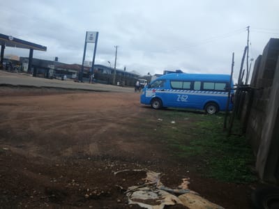 In Ajasse Ipo, Nigeria, a blue passenger van with the markings "Z-62" is parked on a dirt lot next to a paved road. A gas station with a prominent "MRS" sign is visible in the background, along with other buildings and power lines. The sky is overcast, and the ground is covered in dirt and rocks. A person is seen riding a motorcycle in the distance.