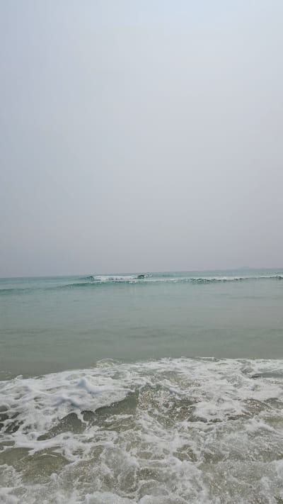 The image shows a sideways view of the coastline at Bac Cam Ranh, Vietnam. The sky is a pale gray, taking up a significant portion of the left side. The sea transitions from a calmer, light green area to an agitated zone of white foamy waves. The waves are closest to the right edge of the frame. The interaction between the water and the beach is visible through the waves. The coastline appears featureless and is in the distance. No people, animals, or man-made objects are clearly visible.