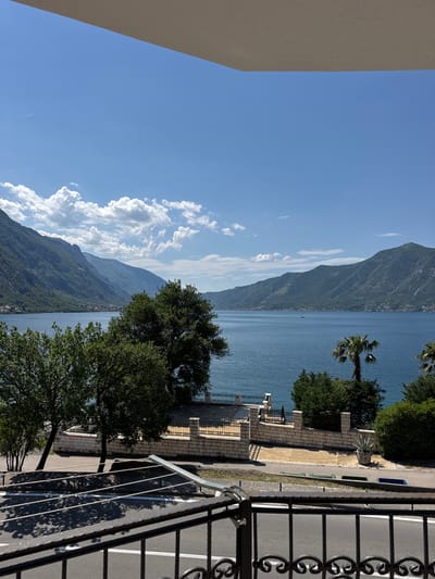 From a high vantage point, likely a balcony, the image captures a panorama in Donji Orahovac, Montenegro. The view includes a clear, blue sky with cumulus clouds to the left, contrasting with towering mountains that descend towards a large body of water. This water, likely the Bay of Kotor, reflects the sky's blue. Closer to the viewer, the scene features vegetation including trees and a palm tree. A low brick wall with black metal railing borders a sidewalk, and beyond that a street. The foreground includes parts of a wrought iron balcony railing. No people are visible in the scene. The environment suggests a bright, sunny day.