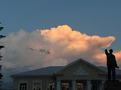 This is a photograph of a monument in Slobozia, Moldova. In the foreground, a dark silhouette of a statue stands on a pedestal, its right arm outstretched and index finger pointing forward. Behind the statue, a building with a pale facade and a grey, corrugated metal roof is visible. The building features classical architectural elements, including columns and decorative pediments above windows. The sky above is a gradient of deep blue at the top, transitioning to lighter hues near the horizon. A large, dramatic cloud formation dominates the upper portion of the image, illuminated by warm, peachy-orange light, suggesting it is either sunrise or sunset. The setting appears to be an open area or square, likely a public space within the city.