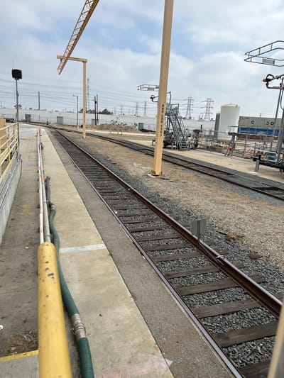 This is a photograph taken from a slightly elevated perspective, looking down at a railway track and adjacent industrial area. The scene is outdoors, under a cloudy sky, suggesting it is daytime. The dominant features are the railway tracks, laid with wooden sleepers and gravel, running diagonally across the frame.
To the left of the tracks, there is a concrete platform with various industrial elements. A tall, yellow metal pole with visible numbering (85565367) stands on the platform, and a section of a yellow crane jib is visible in the upper left corner. Further back, industrial buildings, a large white storage tank, and shipping containers are present. Power lines and transmission towers are prominent in the background, indicating an industrial or utility setting.
On the concrete platform in the foreground, a green ribbed hose and a yellow railing are visible. The presence of hoses and industrial infrastructure suggests some form of operational activity, possibly related to material transfer or maintenance. There are no people visible in the image. The overall impression is one of an active industrial site under overcast skies. The location is identified as Torrance, United States of America.