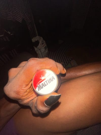 A close-up, dimly lit shot focuses on a hand holding a round, red and white container labeled "MALTINA". The hand, with dark nail polish on one finger, is positioned over the thigh of a person whose skin is visible in the background. The environment appears to be indoors, with a checkered pattern visible on the floor and a striped fabric draped in the background. A white bag with text that includes "MASTERBAKER" is also present. The overall impression is an indoor setting, possibly a room in Akowonjo, Nigeria, with the focus on the person's hand and the beverage container.