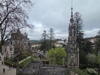 The image displays an elevated view of Quinta da Regaleira in Sintra, Portugal, situated within the civil parishes of Santa Maria e São Miguel, São Martinho e São Pedro de Penaferrim. The scene is set under an uniformly overcast grey sky.
On the left, the multi-storied Regaleira Palace is visible, a grey stone structure exhibiting intricate Neo-Manueline and Gothic Revival architectural elements, including multiple turrets, arched windows, and detailed carvings. A large, leafless deciduous tree partially frames the upper left portion of the palace. To the right, a tall, slender, and ornately carved Gothic-style stone spire, identifiable as the Chapel of the Holy Trinity tower, rises prominently, culminating in a finial and weather vane.
The foreground and midground feature meticulously landscaped gardens with paved pathways, manicured green hedges, and mature trees, including tall conifers. A decorative stone bridge with balustrades connects sections of the estate grounds near the palace. Approximately 20 individuals, dressed in civilian attire, are visible walking casually on the pathways, predominantly near the palace structure.
In the background, beyond the immediate estate, the town of Sintra is discernible, characterized by light-colored buildings with red-tiled roofs interspersed among densely vegetated hills.