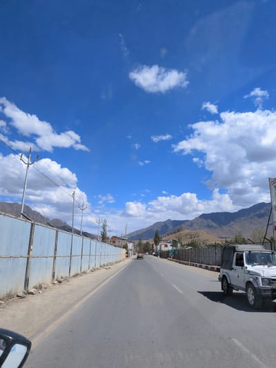 The image depicts a road scene in the city of Brok Khar, India, under a clear blue sky with scattered white clouds. In the foreground, a road stretches into the distance, with a white open-top vehicle parked on the right side. A corrugated metal fence runs along the left side of the road, with utility poles and wires visible above it. In the background, a range of mountains forms the horizon, with some buildings and sparse vegetation dotting the landscape. The overall scene suggests a sunny day with moderate traffic on the road.