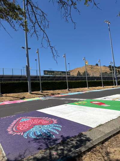 The image shows a parking lot area with painted parking spaces, located at Novato High School in Novato, California, United States of America. The parking spaces are decorated with colorful murals. In the foreground, there is a purple parking space with a heart-shaped design in pink and blue. To the right, a white parking space is visible, adjacent to a green parking space that features an illustration of a ketchup bottle and the letter "S." Further back, a black asphalt road with more painted parking spaces runs parallel to a chain-link fence and bleachers of a sports field. A lush green hedge separates the parking lot from the field. In the background, a grassy hill with trees is visible under a clear blue sky. Several tall light poles are positioned throughout the scene, and a banner with "2021 & 2022 BACK TO BACK STATE CHAMPIONS" is displayed on the bleachers. The overall scene depicts an outdoor environment on a sunny day.
