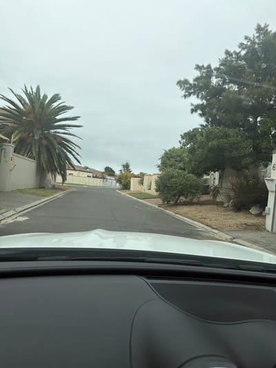 The image appears to be taken from inside a vehicle moving along a street in a residential area. The sky is overcast. A large palm tree is visible on the left side of the road in front of a white building. Various other trees and houses line the street. The number "6" can be seen on a building on the right. Based on the architecture and vegetation visible, the location is consistent with a residential area in Blouberg, South Africa.