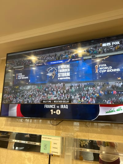 The image shows a large television screen displaying a soccer match broadcast. The screen features graphics related to the "FIFA WORLD CUP 2026" and "PHILADELPHIA." A "SEVERE THUNDER STORM Is approaching" graphic is also prominently displayed, suggesting a potential weather disruption to the event. The match appears to be at halftime, with the score indicating "FRANCE vs IRAQ 1-0." A crowd of spectators is visible in the stadium background, some holding flags. On the right side of the screen, there is a reflection of a person wearing a white shirt and dark pants. Below the television, there's a sign with the text "Good stuff to keep you" followed by options like "[going]," "[refreshed]," and "[cartwheeling]." The overall scene depicts a live sports event potentially impacted by adverse weather conditions.