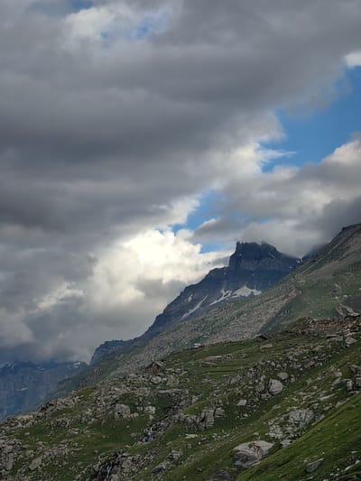 This image depicts a dramatic mountain landscape under a cloudy sky. The foreground is dominated by a rocky, grassy slope, with scattered boulders and patches of green vegetation. A small stream or waterfall is visible cascading down the rocks. In the mid-ground, a steep mountain range rises, characterized by rugged terrain, rocky outcrops, and patches of snow. One prominent peak in the center has a sharp, jagged summit. The upper portion of the image is filled with a heavy, overcast sky, with dark, gray clouds obscuring much of the blue sky, though a few patches of clear blue are visible, along with bright white clouds illuminated by the sun. The overall impression is one of a wild, untamed, and somewhat moody natural environment. The location is identified as Yari-Khoksar, India. There are no people or animals visible in the image.