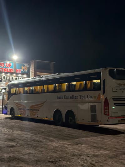 A white bus with "Indo Canadian Tpt. Co." written on its side is parked at night next to a textured, grey surface, possibly a wall or embankment. The bus has illuminated windows, revealing rows of seats inside. A sign for "OR OF RESTAURANT" and "OF DELHI LUNCH" is visible to the left of the bus. Another sign with "INDO-CANADIAN" and a stylized wing graphic is present on the bus. The lighting suggests it is nighttime, with a bright light source to the left casting a glare. The scene appears to be in a commercial or travel-related area, given the restaurant signage and the bus. There are no people clearly visible in the image. The overall atmosphere is dark and somewhat utilitarian.
