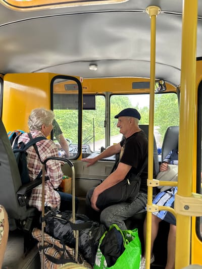 Inside a yellow bus in Pupyshevo, Russia, a female passenger with grey hair, wearing a plaid shirt and a backpack, is seated facing forward. A male passenger, wearing a dark t-shirt and a blue cap, is in the driver's seat, looking forward and smiling. Another passenger, wearing a blue and white patterned garment, is seated to the right of the driver. Luggage, including duffel bags and a green reusable bag, is visible on the floor. The interior of the bus is yellow, with metal poles for support and a window offering a view of green trees outside.