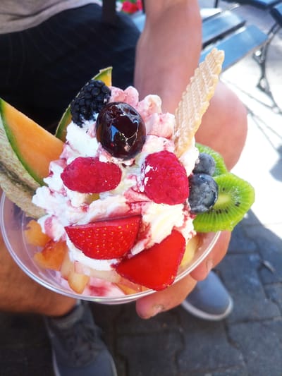 A close-up, high-angle shot shows a person's hands holding a clear plastic cup filled with a colorful fruit parfait. The parfait is topped with whipped cream, a dark red cherry, a blackberry, two raspberries, sliced strawberries, blueberries, kiwi slices, and a decorative wafer cookie. Pieces of melon are visible on the left side of the cup, and diced fruit, possibly peaches, are at the bottom. The whipped cream is drizzled with a red syrup. The background shows the blurred legs and lower torso of a person wearing dark shorts and gray sneakers, as well as a glimpse of a dark bench and some greenery. The scene appears to be outdoors in daylight, possibly a warm day given the attire. The overall impression is a refreshing and appetizing dessert. There is no visible text in the image.