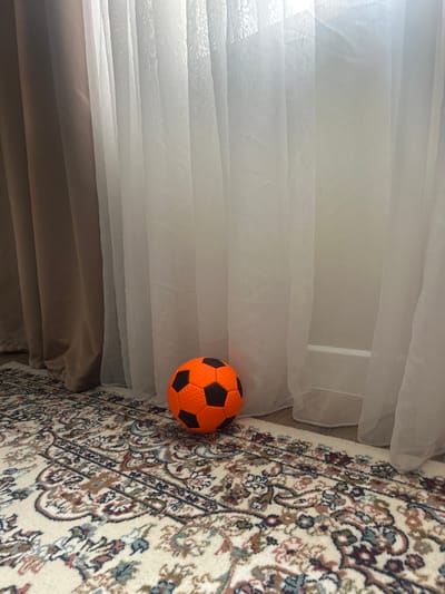 This media file displays an indoor scene, likely a living space, with soft, sheer curtains to the left and a patterned rug to the right. A brightly colored, orange and black soccer ball rests on the floor near the edge of the curtains. The curtains are layered and appear to be diffusing natural light, suggesting it could be daytime. The rug has a detailed, traditional design in cream, red, blue, and brown tones. The overall impression is one of a relaxed, domestic environment. There are no people visible, nor any text. The location context provided is Қырғауылды, Kazakhstan.