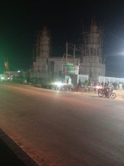 The image depicts a nighttime scene in Tundun Wada West, Nigeria. A large, unfinished structure, likely a mosque given its minaret-like towers and scaffolding, dominates the background. The structure is illuminated by artificial lights, casting a dim glow on the surrounding area.

In the foreground and mid-ground, a street is visible with several motorcycles and bicycles parked or in motion. A few people are gathered around the vehicles, suggesting a social or commercial hub. Some vehicles appear to be trucks or vans. The street itself is paved and dimly lit, with the headlights of vehicles creating streaks of light.

The sky is dark, indicating it is nighttime. The weather appears clear, with no visible signs of rain or fog. The overall atmosphere is somewhat chaotic due to the construction and the presence of numerous vehicles and people.

One notable detail is a large billboard on the left side of the unfinished structure, displaying an image of a man and some text in what appears to be Arabic or a similar script. The text is too blurry to read. Another billboard or sign is visible further to the right, also with unreadable text.

The image seems to capture a moment in the everyday life of Tundun Wada West, highlighting ongoing development and local activity at night.