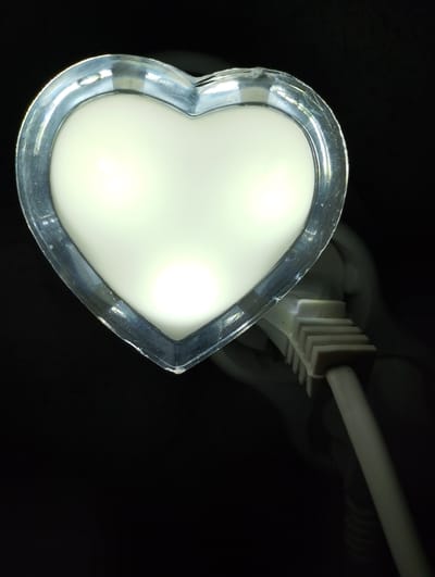 This image shows a close-up of a heart-shaped night light with a transparent outer casing and a frosted white interior. Three bright LED lights are visible within the heart shape, illuminating the interior surface. The night light is plugged into a white electrical cord with a ribbed connector. The background is dark and out of focus, with some light reflecting off the night light's casing. The scene appears to be indoors, likely in a bedroom or child's room, given the nature of the object. There is no text visible in the image. The lighting suggests it might be nighttime or a dimly lit room. The focus is entirely on the night light and its connection to the power source.