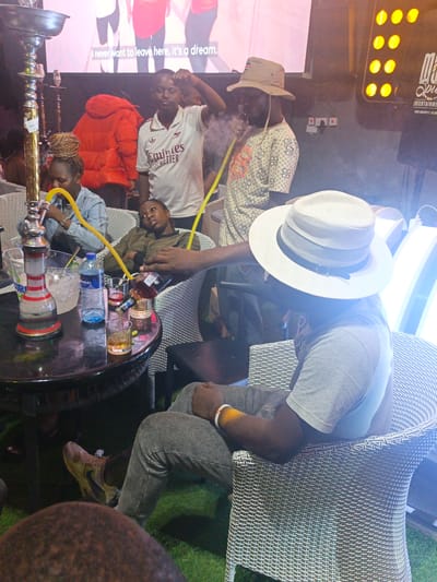 The image depicts a group of people gathered at what appears to be a bar or lounge in Syokimau-Mulolongo ward, Kenya. Several individuals are seated in wicker chairs around a round table, on which there is a hookah, along with drinks including water and what appears to be whiskey. One person is smoking the hookah, while another is pouring whiskey into a glass. Several other people are seated around the table, and a man in an Arsenal jersey is standing nearby. In the background, there is a screen displaying text and some lighting, and a logo for an entertainment company "Spice Entertainment" can be seen on a light board. The flooring is artificial grass.