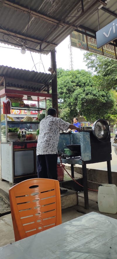 The image captures a scene at an outdoor food stall in Langnibong, Indonesia. A man in a patterned shirt is actively preparing food on a stove. Behind him, another man in a blue shirt is visible. The food stall itself is equipped with shelves displaying various ingredients, including vegetables and packaged goods. A large metal basin sits on a stand to the right of the stove. In the foreground, an orange plastic chair is positioned near a table covered with a white sheet. The environment appears to be a roadside setting, with a corrugated metal roof providing shade and a backdrop of lush green trees. A white plastic container is situated on the ground to the right. The overall impression is that of a bustling local eatery.