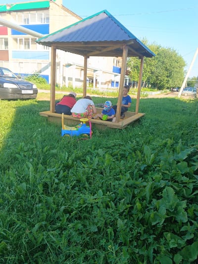 In this outdoor daytime scene, likely in Tolbazy, Russia, a wooden gazebo with a dark blue metal roof stands over a sandbox. Three children are present: two girls and one boy. The two girls are seated in the sandbox, one wearing a red shirt and the other a white shirt with brightly patterned leggings. The boy, wearing a blue t-shirt and a baseball cap, is also in the sandbox, holding a green watering can. A blue toy truck with a yellow handle is situated near the children in the sandbox.
In the background, a multi-story building with blue and beige siding is visible, along with a black car parked on the left side of the frame, displaying the license plate "x302cx 102". The area surrounding the sandbox is covered in lush green grass and various plants. Trees line the background, and the sky above is clear blue, suggesting fair weather. The overall atmosphere appears relaxed and leisurely, indicative of children playing outdoors on a pleasant day.