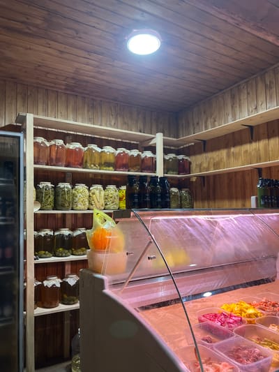 The image depicts shelves filled with jars of preserved foods, likely in a shop or pantry in Pitsunda, Abkhazia. The shelves are constructed of light-colored wood, contrasting with the darker wooden plank walls and ceiling. A bright ceiling light illuminates the scene. On the shelves, numerous glass jars of various sizes contain preserved fruits, vegetables, and possibly olives. Some jars are filled with amber or reddish liquids, while others contain green or yellow contents. There are also several dark-colored bottles on the middle shelf. In the foreground, a refrigerated display case is visible, containing plastic containers filled with various food items, including what appear to be pickled vegetables and seafood. The overall impression is one of a well-stocked establishment offering preserved goods.