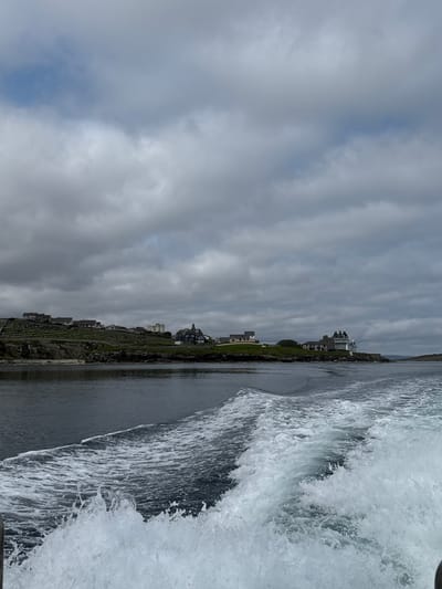 This image captures a view from a boat moving through choppy, dark water, creating a prominent wake in the foreground. The wake is a spray of white foam and splashing water. In the middle ground, a rocky coastline is visible, topped with a scattering of buildings and greenery. A cluster of houses with white roofs lines the upper part of the slope, with some appearing to be terraced. Further along the coast, a larger, white building with multiple windows and a prominent tower stands out. The sky above is overcast with heavy grey clouds, with hints of blue sky peeking through in the upper left portion of the image. The overall impression is a somewhat somber, cloudy day at sea. The scene depicts a coastal settlement, likely a town or village, viewed from a body of water. The location in the city of Lerwick, United Kingdom, is not explicitly identifiable from the visual information provided in the image itself; it requires external context to make such a determination.