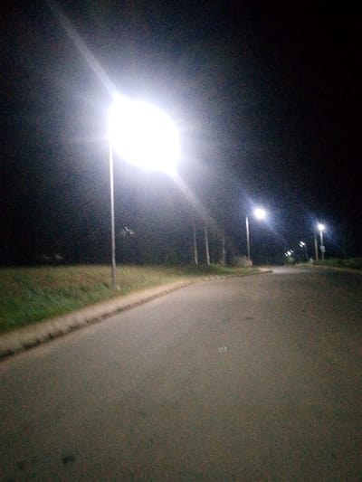A nighttime road is illuminated by a series of streetlights, casting bright glows along the asphalt surface. The closest light is intense, almost blinding, while others appear diminished in the distance. A grassy area borders one side of the road, contrasting with the dark background. The scene conveys a sense of urban isolation, punctuated by artificial illumination.
