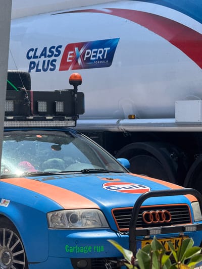 The image shows a close-up view of a blue and orange Gulf-branded vehicle, possibly a truck or van, parked next to a white vehicle with red and blue stripes. The white vehicle has "CLASS EXPERT FORMULA" written on its side. The Gulf vehicle has the iconic Gulf logo and "Gulf" written in white within an orange circle. It also has a grille with four orange rings, resembling the Audi logo.

The front of the Gulf vehicle is visible, including its headlights and a license plate that reads "Y-K". The vehicle appears to be custom-painted with racing stripes. The side of the vehicle has green text that reads "Garbage run". There are also some lights attached to the front of the Gulf vehicle.

The scene appears to be outdoors, possibly in a parking lot or industrial area, given the presence of the two vehicles and the lighting equipment. The lighting suggests it is daytime, and the weather appears to be clear. There are some green leaves from a plant obscuring the top right corner of the image. The image is taken from a low angle, looking up at the vehicles. The overall impression is of a commercial or service vehicle.

The location context is Liješnje, Montenegro.