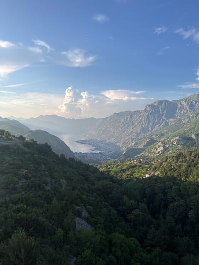 A scenic, high-angle view captures the Bay of Kotor in Čavori, Montenegro, under a bright, partly cloudy sky. In the foreground, dense green forest blankets steep hillsides, with scattered grey rocks and trees visible. Further back, the bay stretches out, with a town nestled along its shores, its buildings appearing as a light-colored cluster. The bay is surrounded by dramatic, rugged mountains that rise sharply, their slopes covered in a mix of vegetation and rocky outcrops. Sunlight streams in from the left, casting shadows and highlighting the terrain. In the distance, the mountains fade into a hazy, atmospheric perspective. A few scattered clouds drift across the blue sky, one particularly large, puffy cloud stands out near the horizon. The overall scene evokes a sense of natural beauty and tranquility.