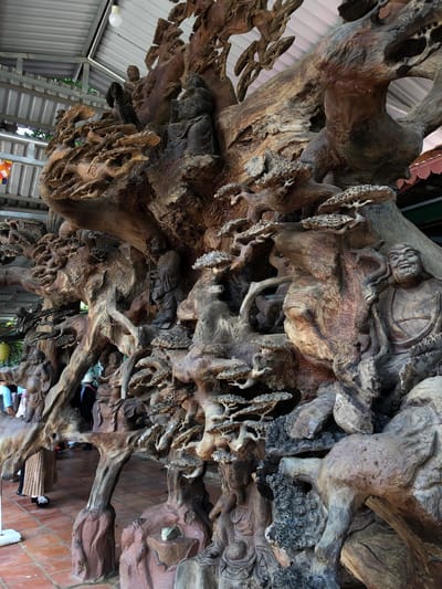 This image captures a large, intricate wooden sculpture, likely a centerpiece within a temple or a sacred space in Nam Ban, Vietnam. The sculpture is a complex arrangement of gnarled wood, possibly depicting a scene from Buddhist mythology. Several carved figures, seemingly monks or deities, are integrated into the naturalistic forms of the wood. Some figures are seated in meditation, while others stand, holding staffs or in prayerful poses. The wood itself is textured and aged, with a rich brown patina that highlights the carvings. The sculpture is partially sheltered by a corrugated metal roof, suggesting it is outdoors or in an open-air pavilion. The ground is paved with terracotta tiles. In the background, several people are visible, some appearing to be visitors observing the artwork, while others might be residents or temple staff. The overall impression is one of a spiritual and artistic environment, deeply rooted in local culture and tradition.