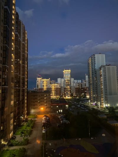 The image captures a twilight view of a residential complex in Korkino, Russia. On the left, a tall, modern apartment building with numerous balconies and illuminated windows dominates the foreground. To the right, a cityscape unfolds with several other high-rise buildings, some featuring decorative lighting on their upper floors. A network of roads with parked cars is visible, and streetlights illuminate the paved areas. The sky above is a deep blue, transitioning to darker tones, with a few scattered clouds. Green spaces with trees and benches are interspersed between the buildings. The overall impression is that of a developing urban area at dusk.