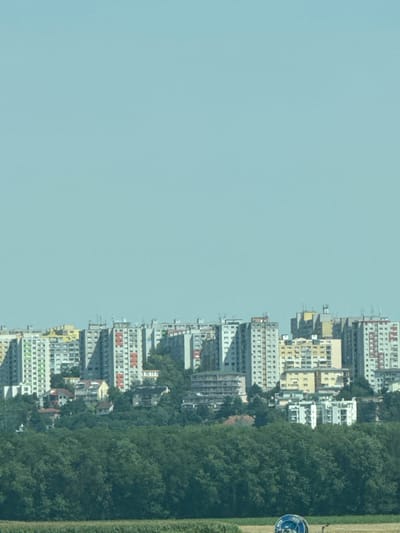 The image captures a wide view of a residential area in Wolfsthal, Austria, under a clear, pale blue sky. In the foreground, a dense, dark green forest stretches across the lower portion of the frame. Beyond the trees, a collection of high-rise apartment buildings dominates the midground and background. These buildings are diverse in height and architectural style, with many featuring repetitive window patterns and balconies. Some of the buildings are painted in muted tones of gray, white, and yellow, while others have accents of red and green. Smaller houses and buildings are nestled among the trees and at the base of the apartment blocks. In the far distance, the sky is a uniform, light blue. At the bottom of the frame, a blurred field is visible, and a small portion of a blue and white circular object, possibly a piece of machinery or signage, is present in the lower right corner. The scene appears to be a snapshot of urban and natural landscapes coexisting.
