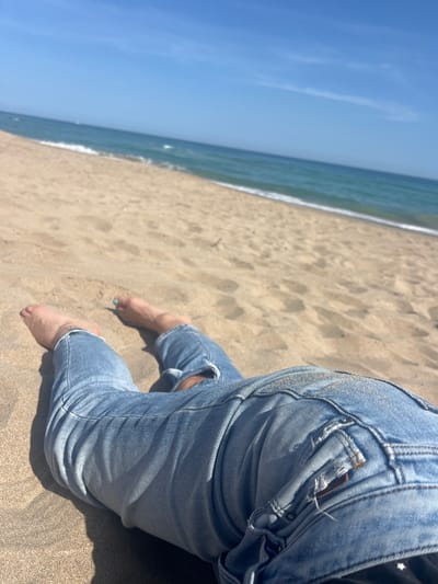 This bright, sunlit photograph captures a person's lower body as they sit or kneel on a sandy beach, facing a calm sea under a clear blue sky. The person is wearing light blue denim jeans, which show some wear and tear with rips around the knees. Their bare feet are visible in the golden sand, with one big toe displaying light blue or turquoise nail polish. Strong sunlight casts shadows from the person onto the sand, indicating it is likely midday or early afternoon on a clear, sunny day.
The scene depicts a peaceful and relaxed atmosphere at a beach location, consistent with La Marina De Creixell, Spain. The ocean waves are gently breaking near the shore, marked by a thin line of white foam. The vast expanse of the clear blue sky dominates the upper left portion of the image, while the sandy beach fills the foreground and midground, leading towards the water. The composition is taken from a low angle, with the horizon slightly tilted, offering a casual perspective of a tranquil beach day. There is no visible text in the image.