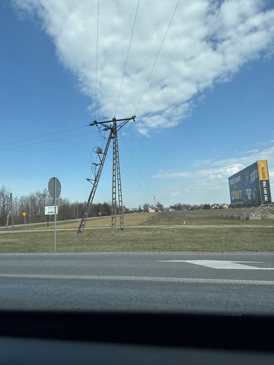 The image depicts an outdoor, daytime scene in Łomna-Las, Poland, under a partly cloudy blue sky. The foreground shows an asphalt road surface with a double white line on the right side and a prominent white arrow painted on the pavement, indicating a turn to the right. A dark, out-of-focus band occupies the bottom portion, suggesting a view from a vehicle.
In the midground, a tall, brown-grey lattice-work electricity pylon, constructed with an A-frame base, stands in a wide, dormant grassy field to the right of the road. Multiple power lines extend horizontally and diagonally from the pylon's cross-arms. To the left of the pylon, adjacent to the road, a circular regulatory road sign on a pole is partially visible, with a smaller white rectangular sign attached below it. Further left, a yellow triangular warning sign is also present.
Behind the pylon and signs, a line of bare deciduous trees forms a sparse treeline. Further to the right in the midground, a large rectangular billboard advertising "LVBET" with a phone number "791 301 708" is positioned within the field. In the far distance, across the field, several houses with light-colored roofs are visible, scattered among trees, indicating a rural settlement. No people or active interactions are observed in the scene.